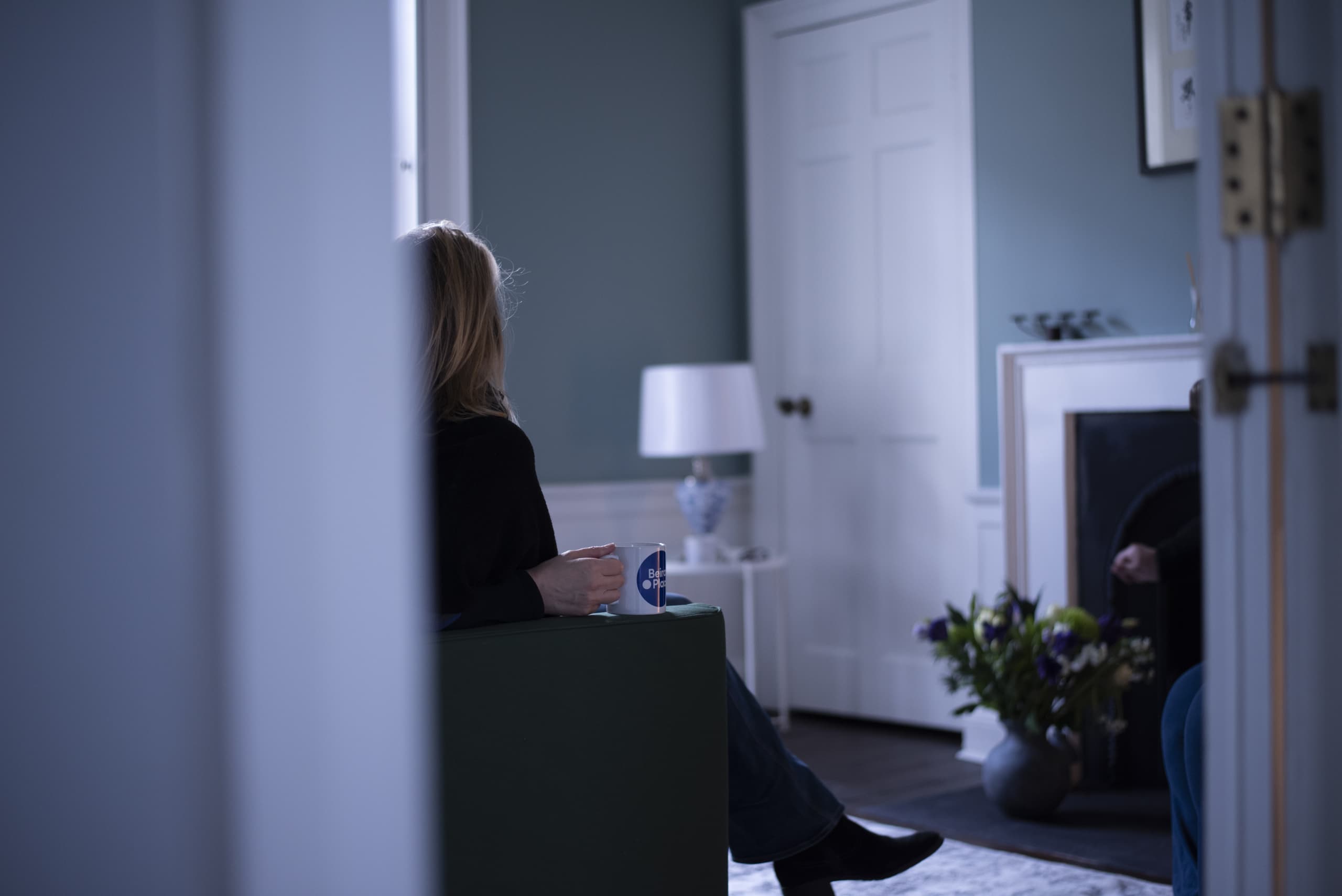 A woman holding a 'Beira's Place' mug in a cozy room with muted green walls, a fireplace, and flowers.